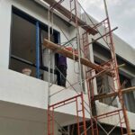 A two-story modern tropical villa under construction in Bali, showing scaffolding, window frame installation, and white exterior wall finishing..