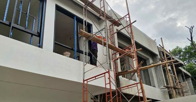 A two-story modern tropical villa under construction in Bali, showing scaffolding, window frame installation, and white exterior wall finishing..